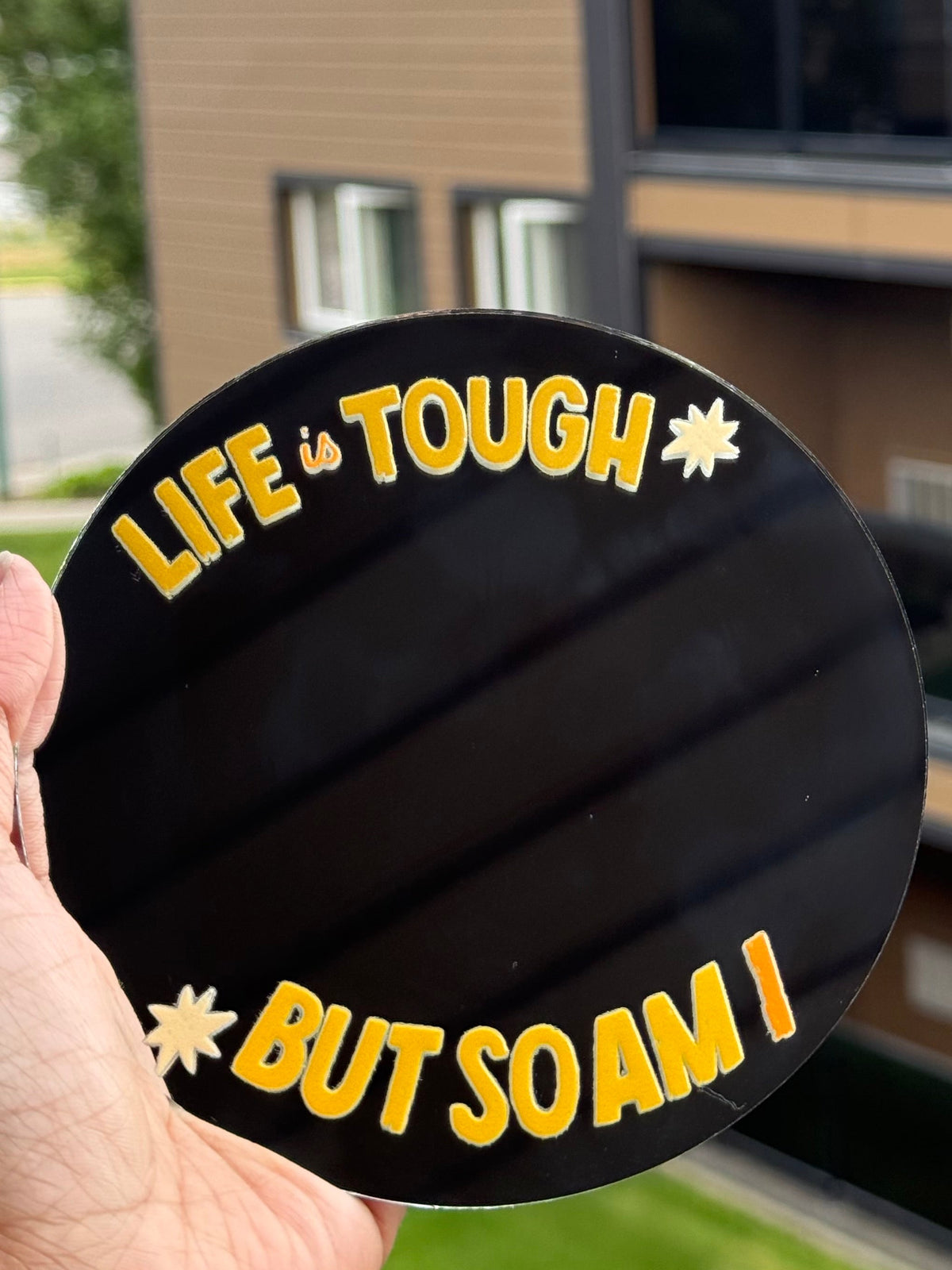 A hand holds a round black sticker or coaster in front of a blurred apartment building. The sticker has yellow text reading 'LIFE IS TOUGH BUT SO AM I' with small yellow starburst accents.
