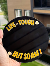 A hand holds a round black sticker or coaster in front of a blurred apartment building. The sticker has yellow text reading 'LIFE IS TOUGH BUT SO AM I' with small yellow starburst accents.